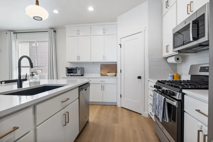 White cabinetry with bronze-finish hardware, stainless steel appliances, wood-finish flooring, white countertop surfaces, and a kitchen island with an undermount sink and matte black faucet