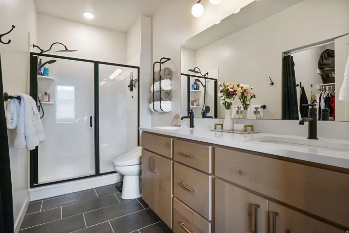 Dual vanity with integrated sinks and white countertops, wood-finish cabinetry, black fixtures, glass-enclosed shower with black framing, and gray tile flooring