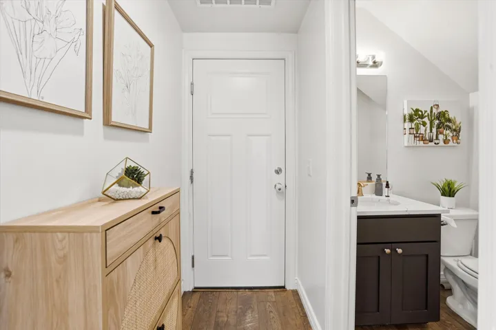 Wood-finish flooring throughout, white paneled door, dark wood vanity with white countertop, wall-mounted light fixture, and white toilet
