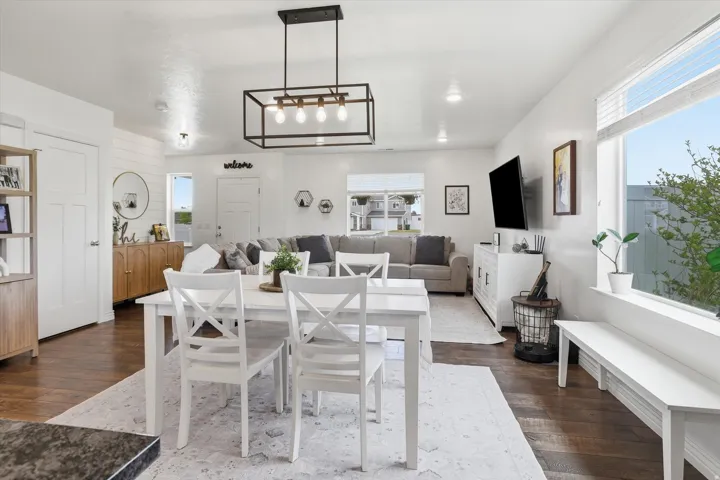 Open-concept living area featuring a wood-finish plank floor, white shaker-style dining chairs with a light wood table, recessed ceiling lighting, a modern rectangular pendant fixture, and abundant natural light from large windows