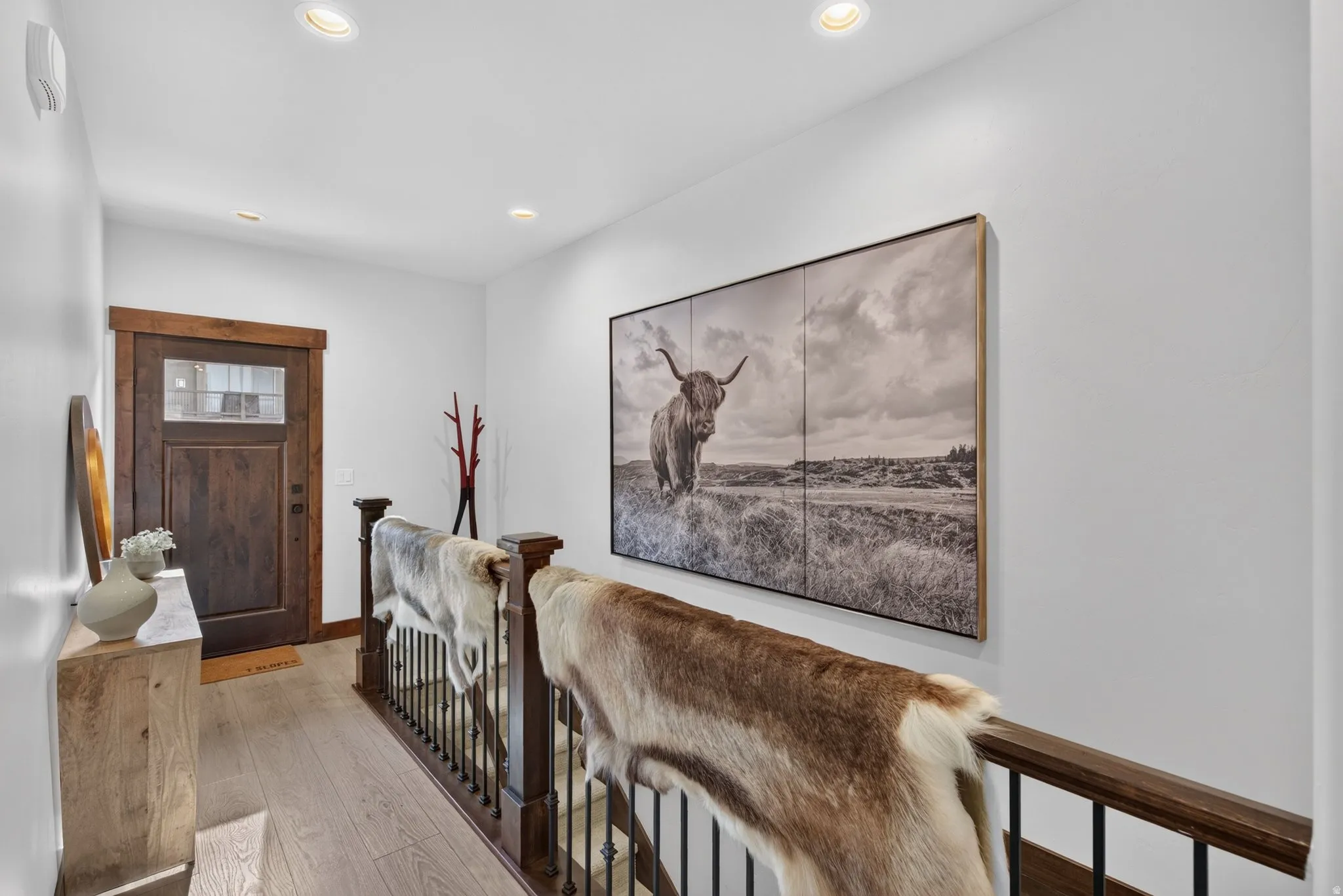 Recessed ceiling lighting, wood-finish flooring, dark wood entry door with glass panel, dark wood railing with black spindles, and a light wood-finish console table