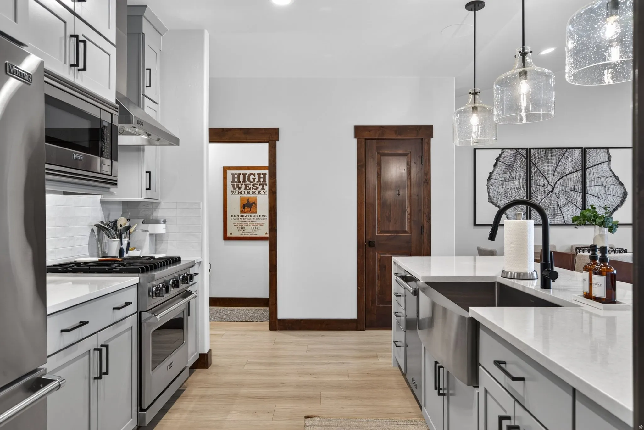 Light wood-finish flooring, light gray cabinetry with black hardware, stainless steel appliances, white subway tile backsplash, and clear glass pendant lights