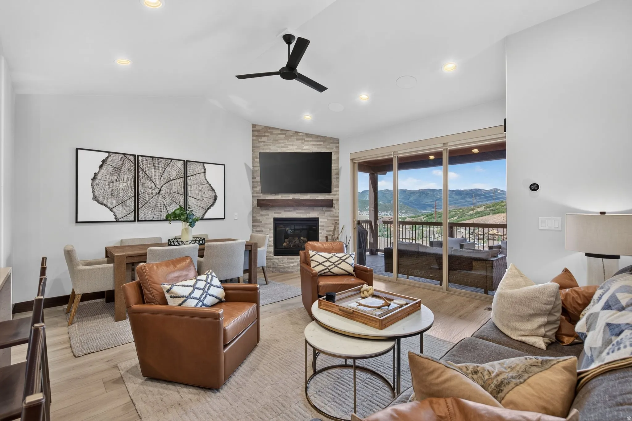 Vaulted ceilings with recessed lighting, stone-clad fireplace with wood mantel, wood-finish flooring, and sliding glass doors opening to an outdoor deck