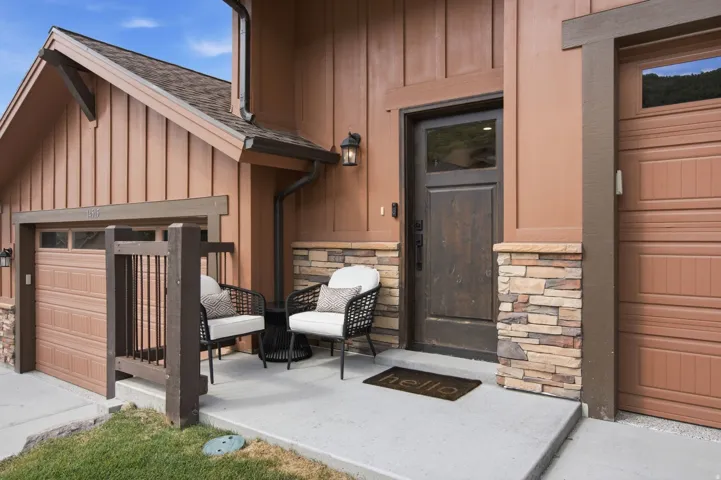 Board-and-batten siding with stone veneer accents, concrete porch with wood railing, dark wood entry door with frosted glass, and two garage doors