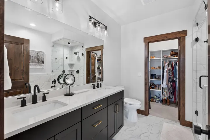 Dual vanity with black cabinetry and gold-tone hardware, white countertop, walk-in shower with glass enclosure, white marble-style flooring, and walk-in closet with built-in shelving