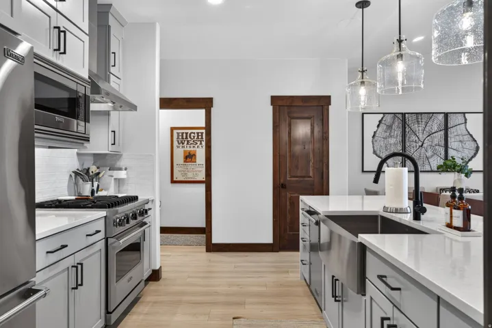 Light wood-finish flooring, light gray cabinetry with black hardware, stainless steel appliances, white subway tile backsplash, and clear glass pendant lights
