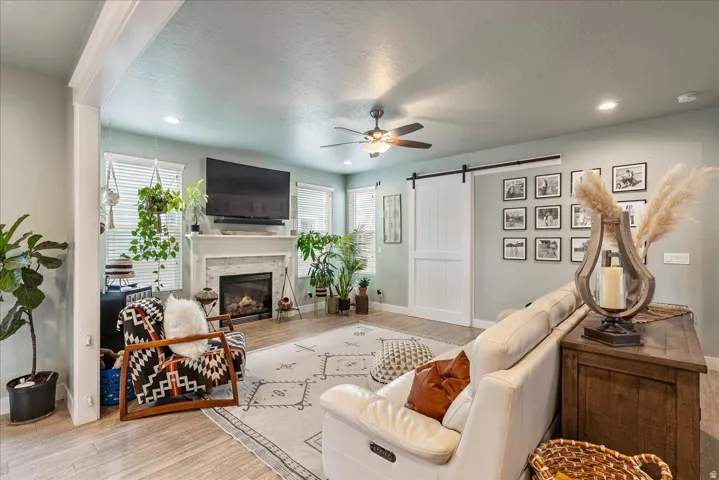 Spacious living area featuring light wood-finish flooring, a white mantel fireplace with stone surround, recessed lighting, and a ceiling fan