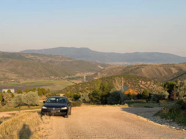 Expansive mountain scenery with a distant valley, and a cabin-style structure among varied trees