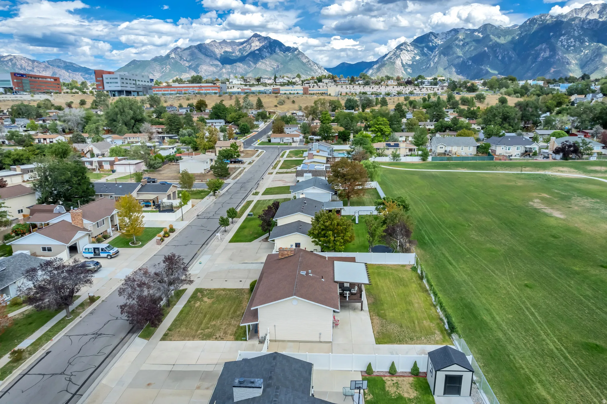 Expansive mountain range backdrop, residential neighborhood with paved streets, adjacent to a large grassy field, and a property featuring a separate outdoor storage shed