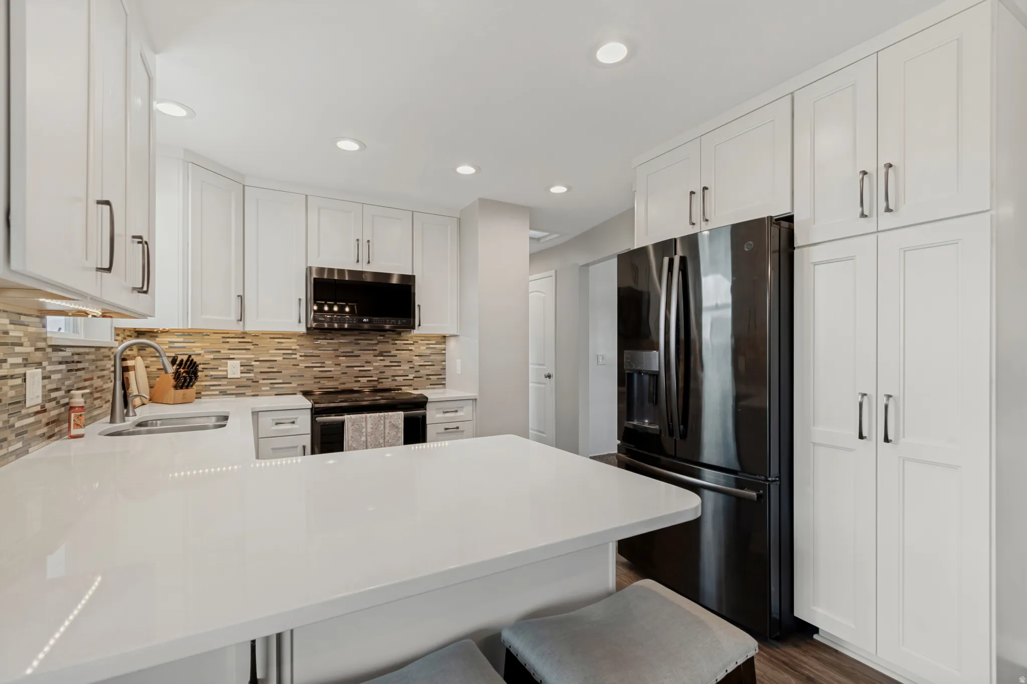 White shaker-style cabinetry, solid surface countertops, mosaic tile backsplash, black stainless steel appliances, and recessed lighting