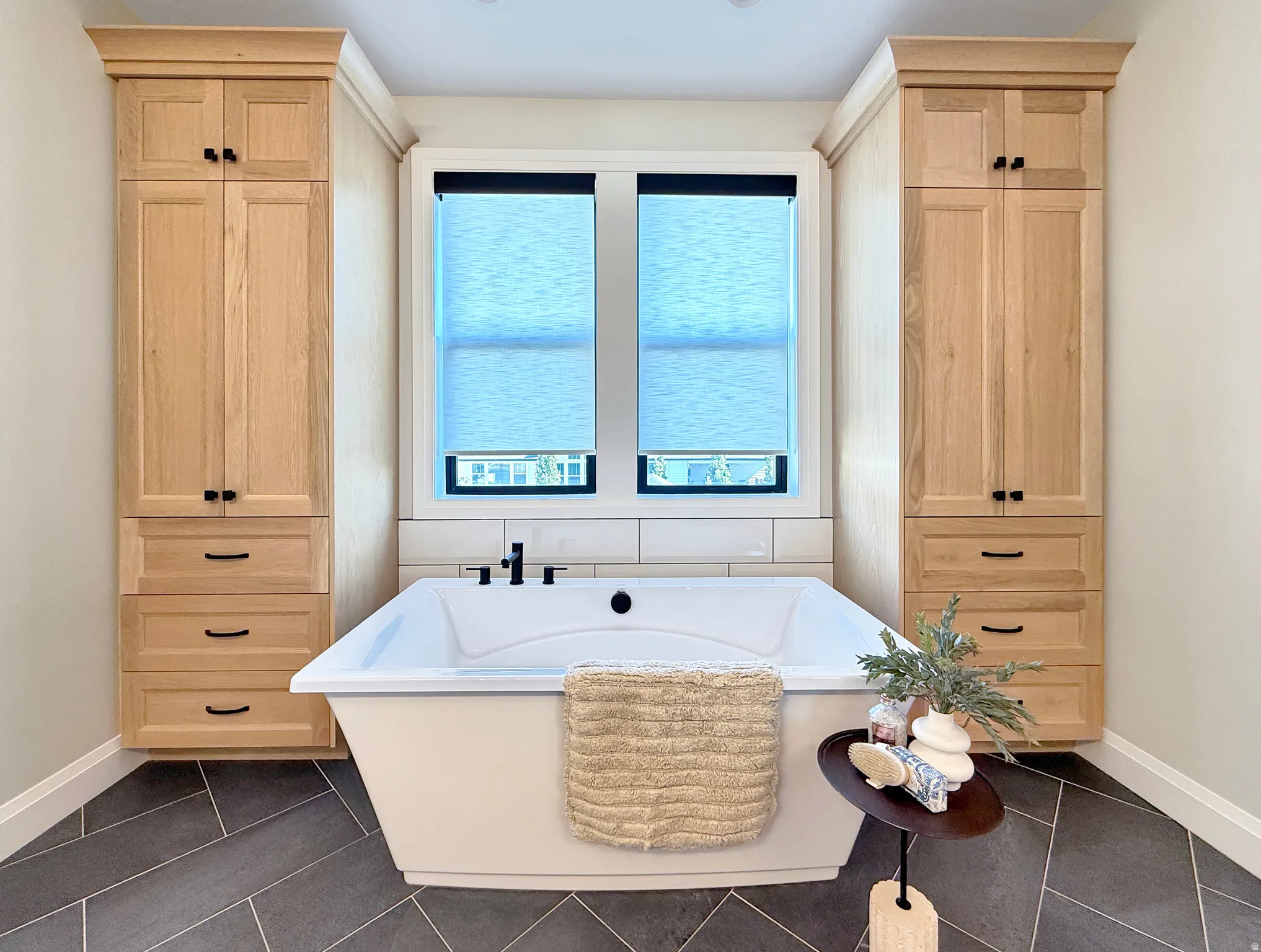 Freestanding soaking tub with matte black fixtures, flanked by built-in wood-finish cabinetry, under windows with roller shades, set on herringbone-patterned tile flooring