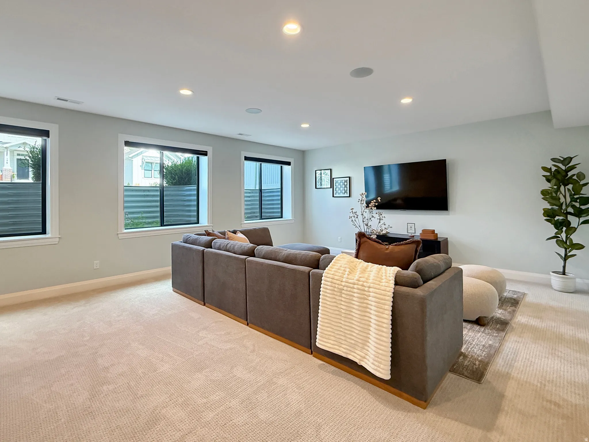 Recessed lighting, wall-mounted television, light-colored carpeting, three windows with white trim, and a built-in ceiling speaker