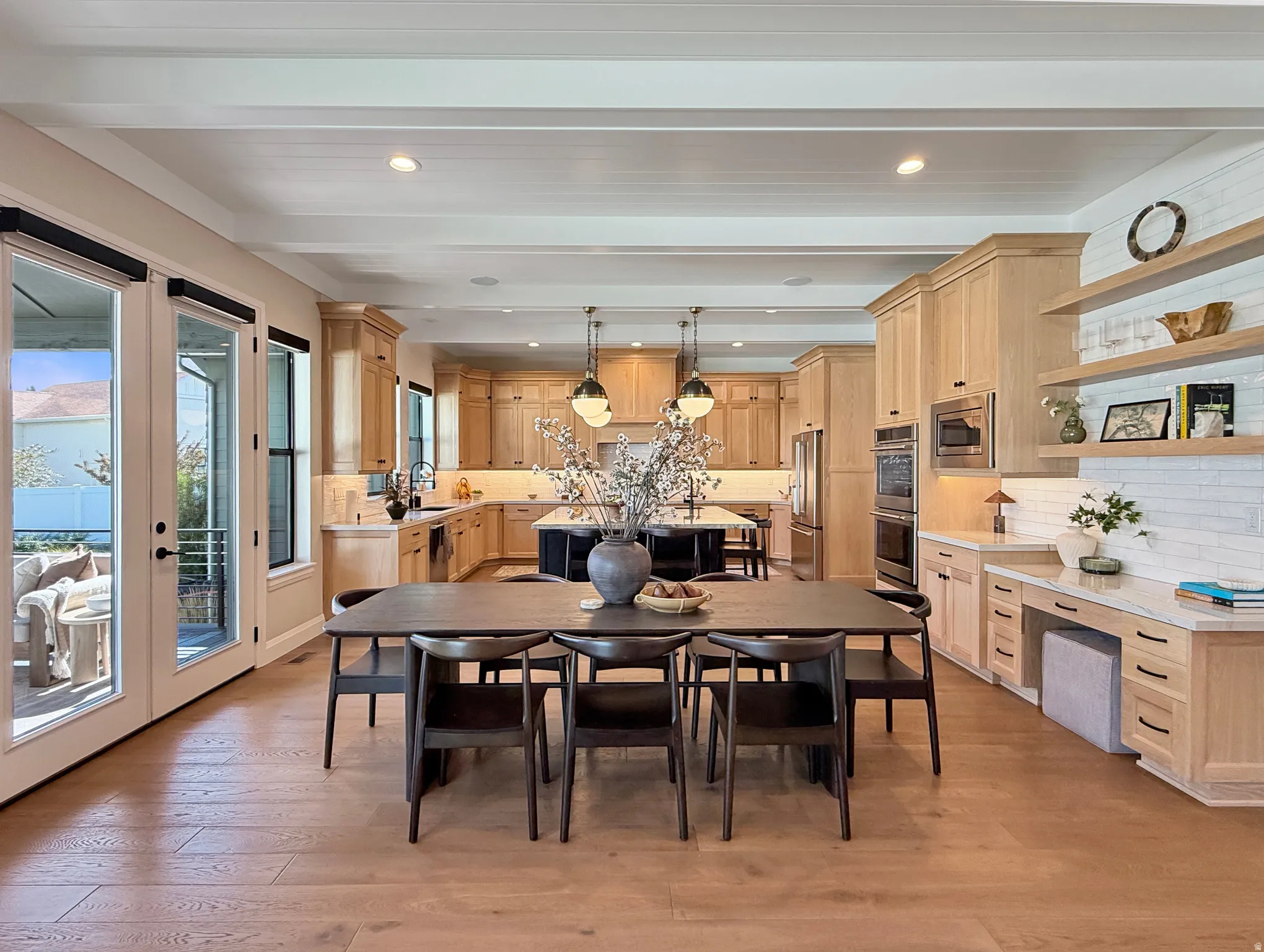 Expansive open-concept living area featuring light wood-finish flooring, a kitchen with light-toned cabinetry, a built-in desk area with open shelving, and double glass-paneled doors