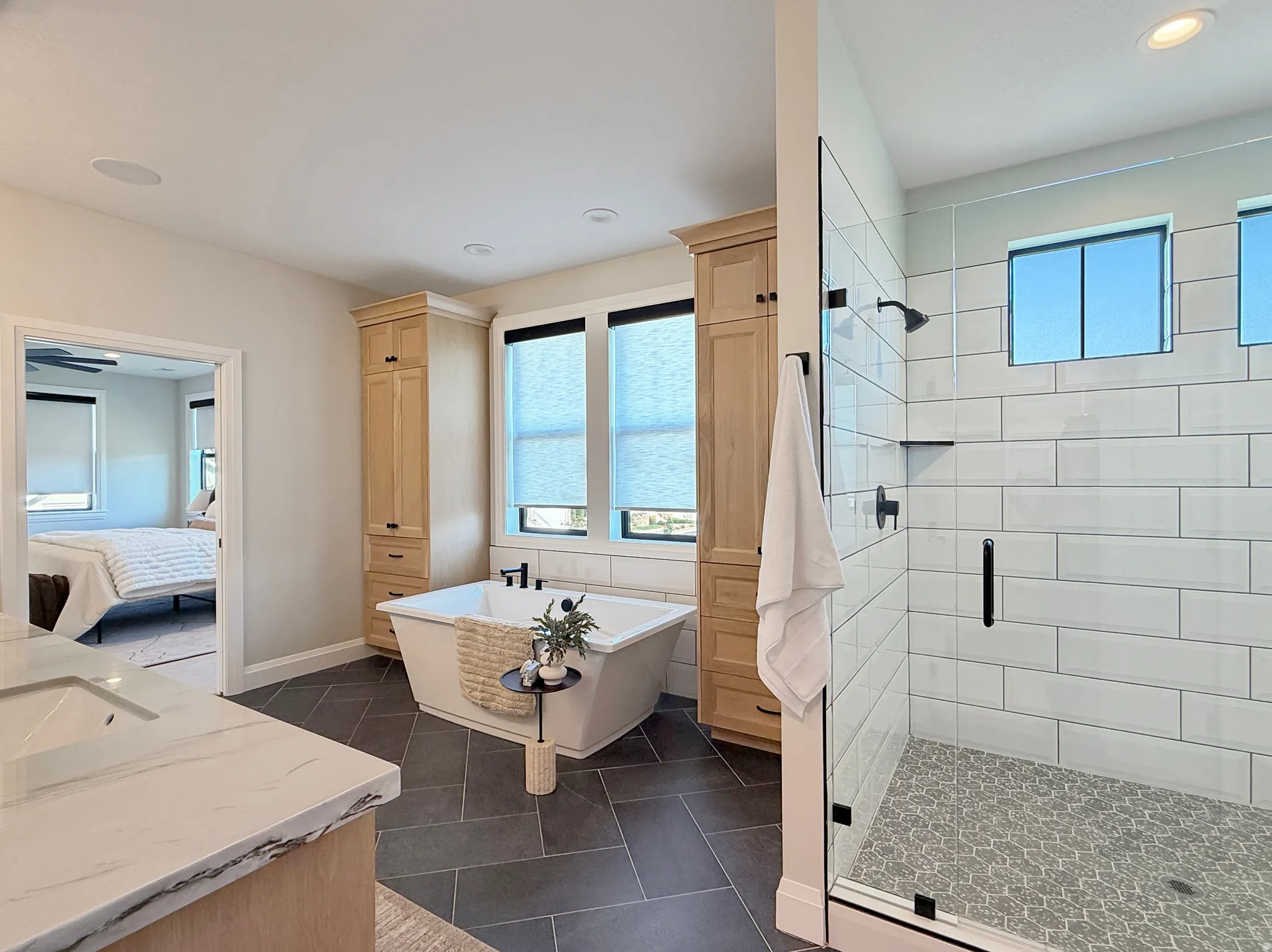 Freestanding soaking tub, floor-to-ceiling cabinetry, glass-enclosed shower with subway tile surround, patterned shower flooring, and dual rectangular windows