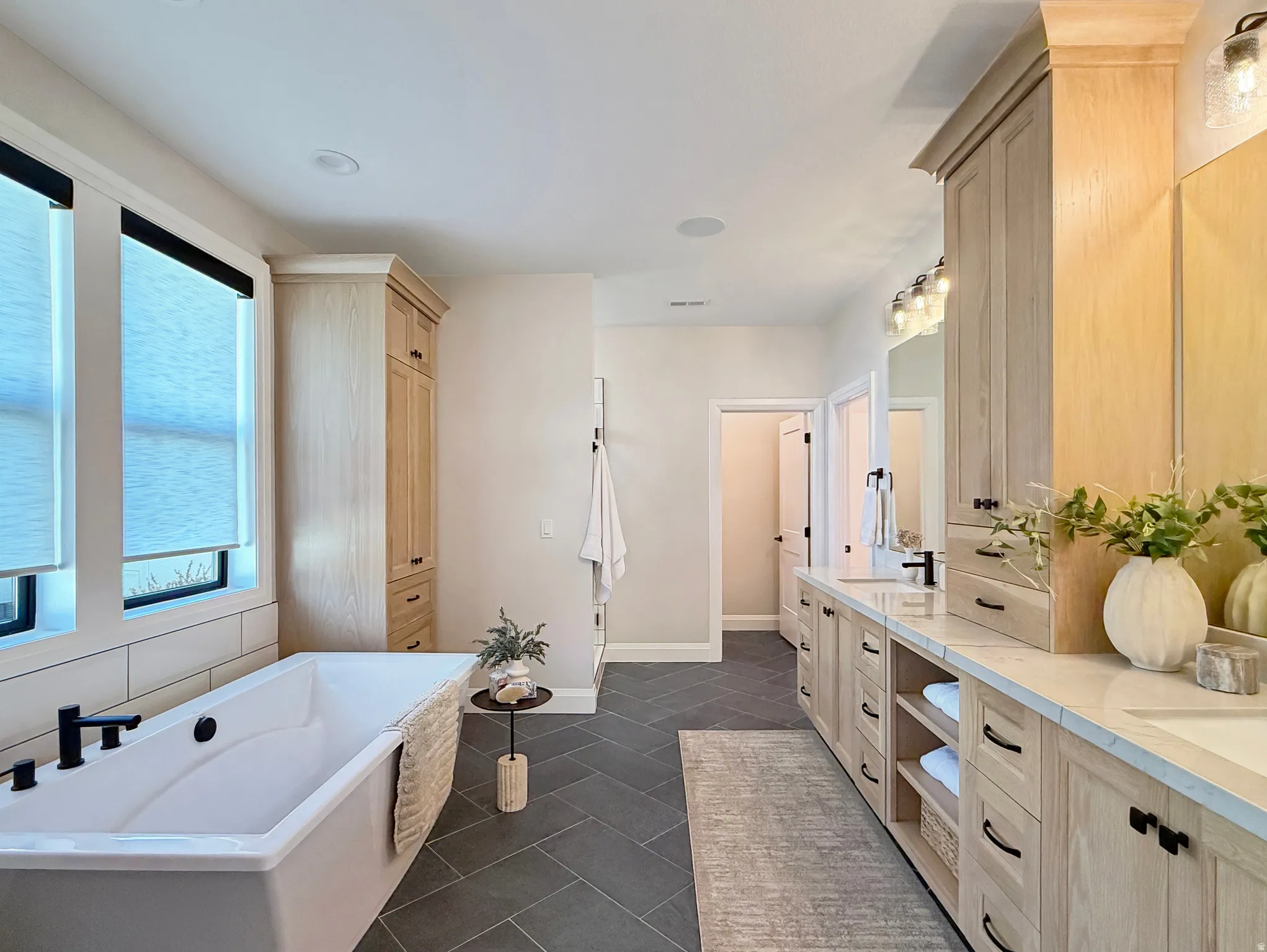 Freestanding soaking tub with matte black fixtures, light wood-finish cabinetry with black hardware, double vanity with integrated sinks, and herringbone-patterned floor tile