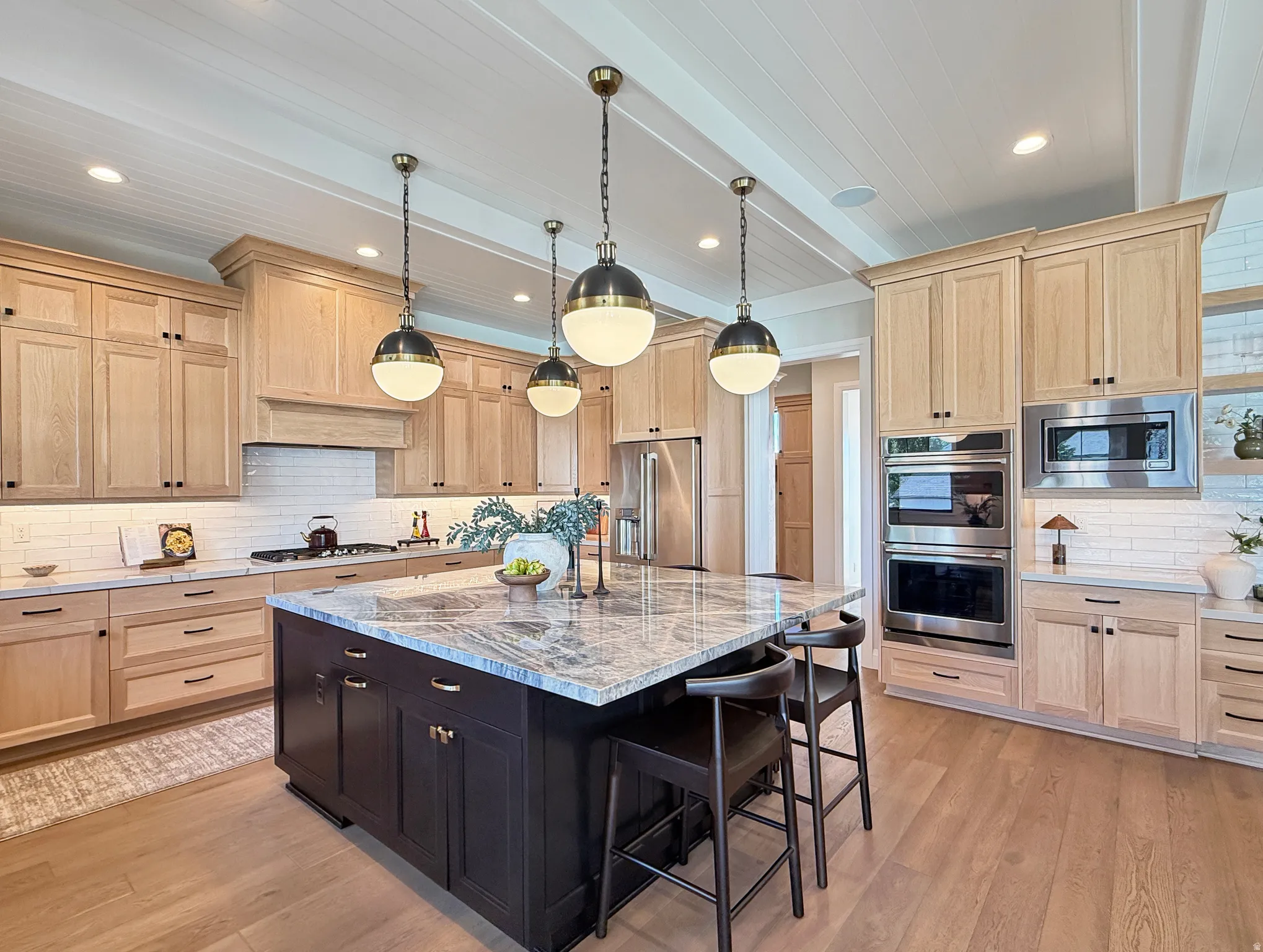 Kitchen featuring light wood-finish cabinetry, a central island with a stone countertop, an integrated stainless steel oven and microwave, a tile backsplash, and wood-finish flooring