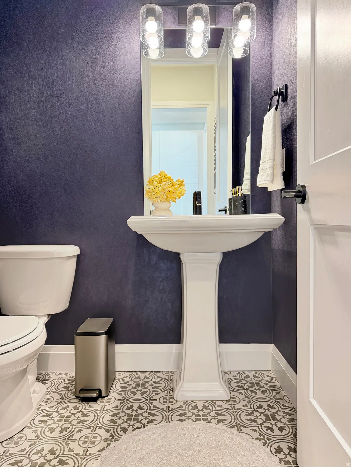 Pedestal sink with a framed mirror, three-light vanity fixture, patterned floor tiling, and a white paneled door