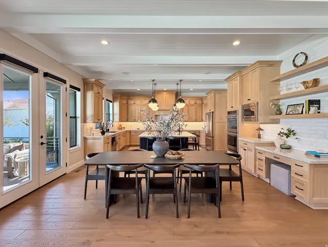 Expansive open-concept living area featuring light wood-finish flooring, a kitchen with light-toned cabinetry, a built-in desk area with open shelving, and double glass-paneled doors