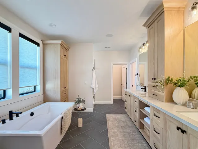 Freestanding soaking tub with matte black fixtures, light wood-finish cabinetry with black hardware, double vanity with integrated sinks, and herringbone-patterned floor tile