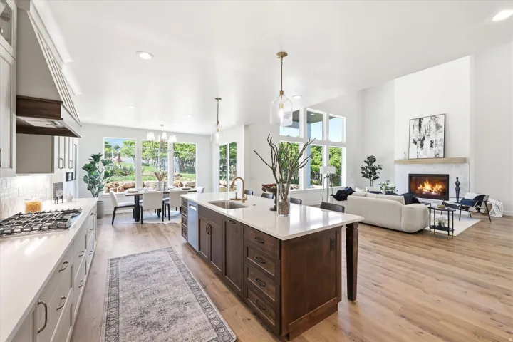 Open-concept interior featuring light wood-finish flooring, a kitchen island with a built-in sink, a gas cooktop with an overhead range hood, a dining area with expansive windows, and a living area with a fireplace