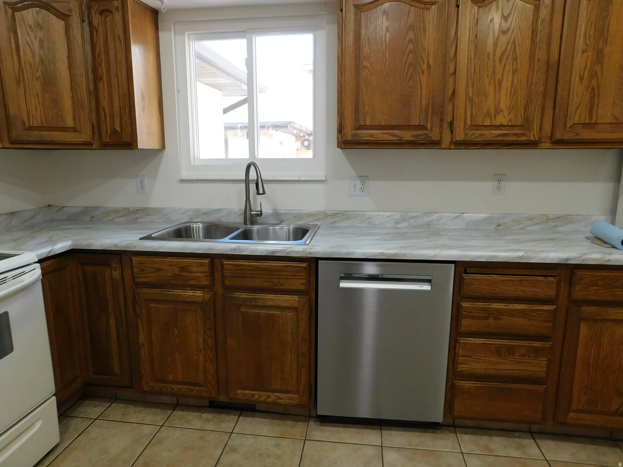 Kitchen featuring wood cabinetry, light-toned countertops, a stainless steel dishwasher, a double basin sink, and tile flooring