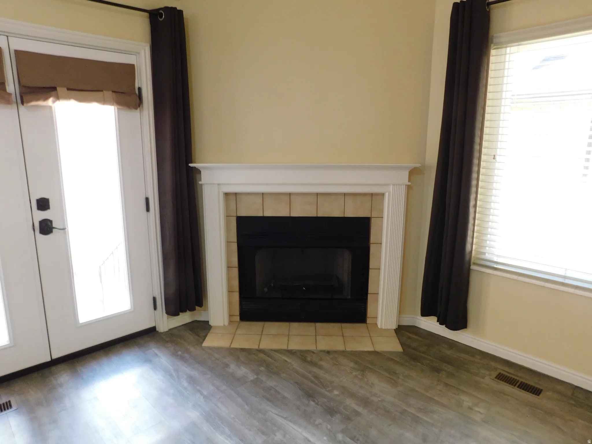 Fireplace with tile surround and white mantel, wood-finish flooring, and French doors with privacy shades