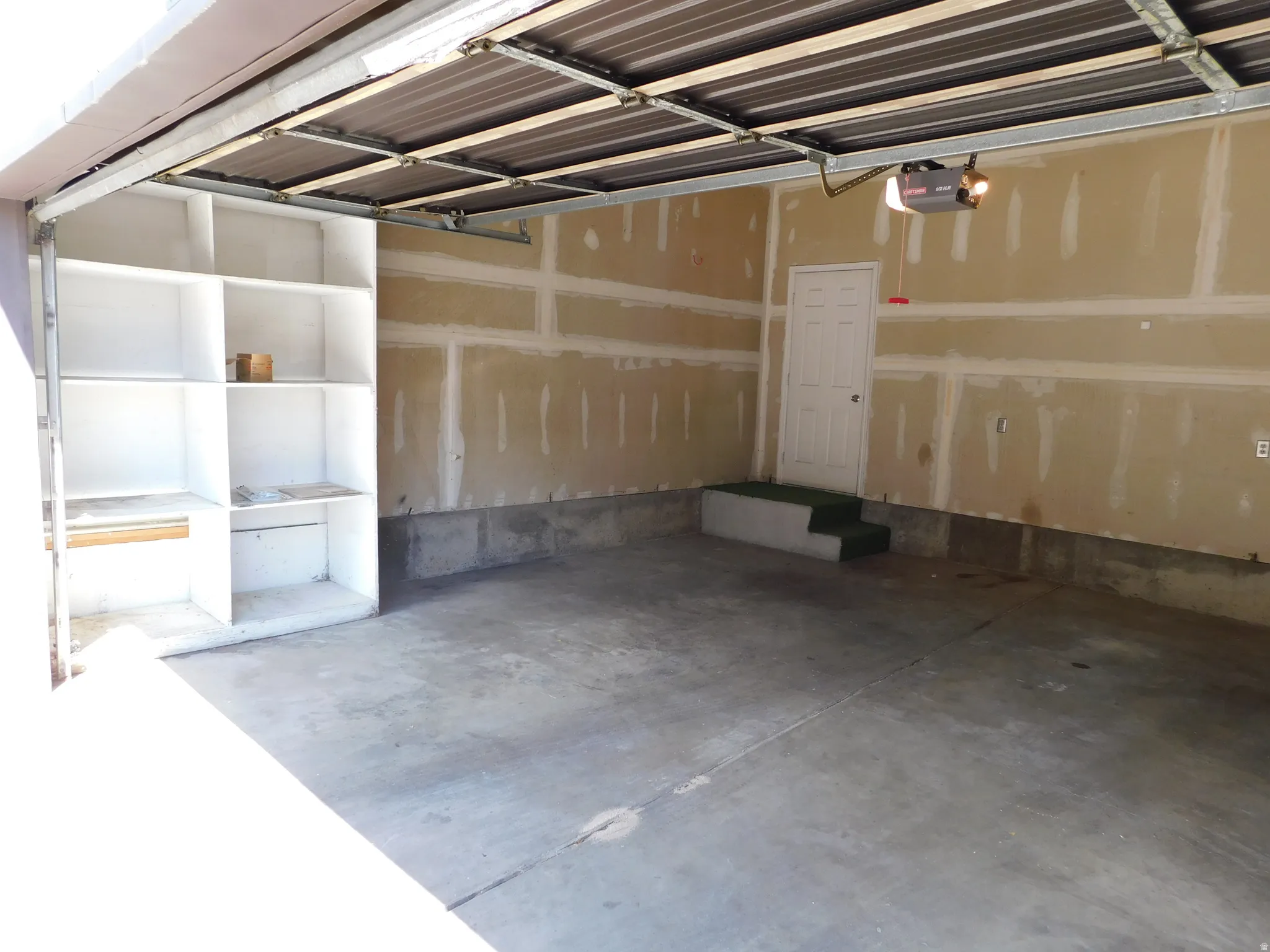 Concrete flooring, built-in shelving, white entry door, steps with green carpeting, and an automatic garage door opener