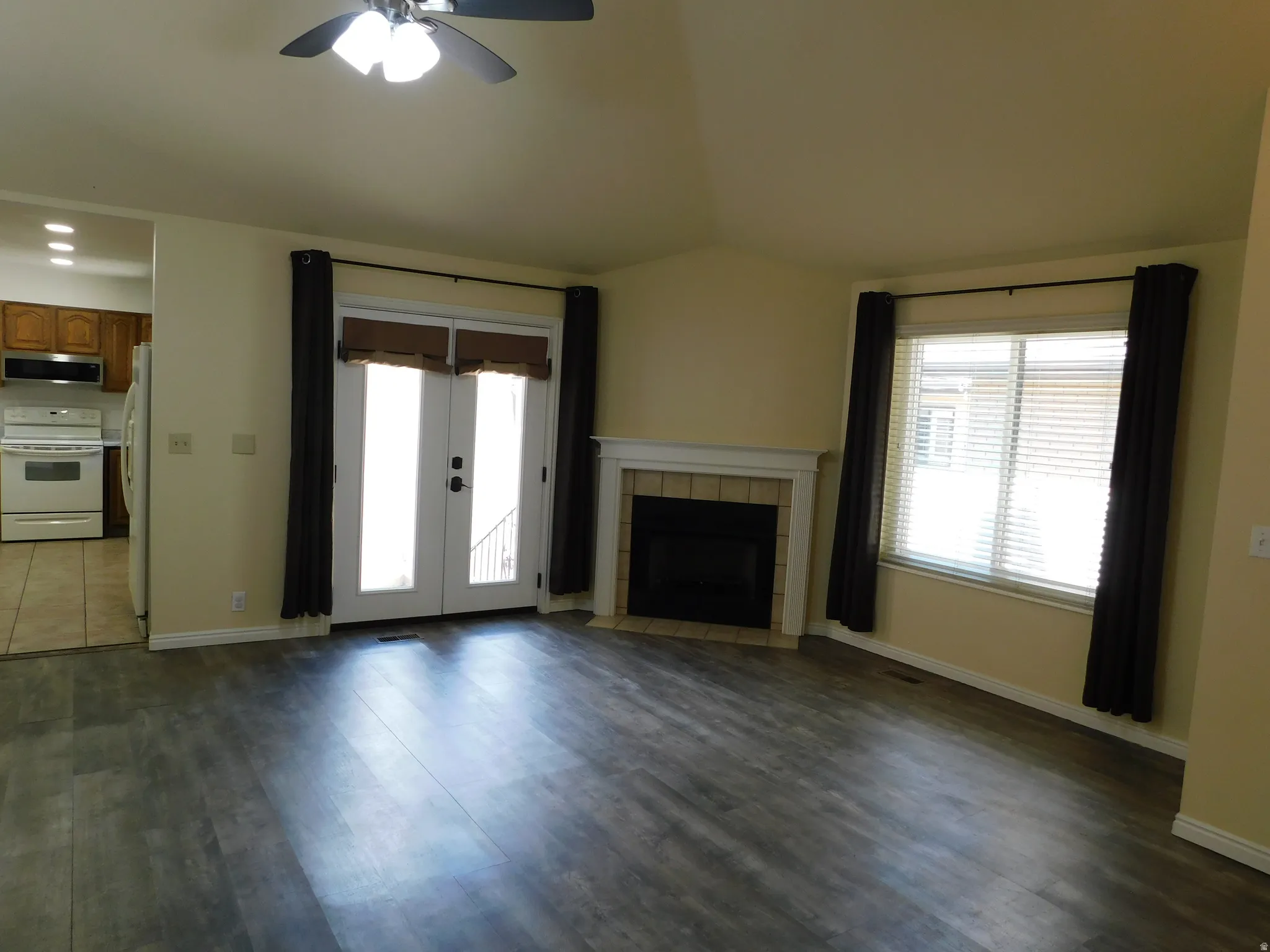 Wood-finish flooring, corner fireplace with white mantel, white French doors, ceiling fan with integrated lighting, and a large window