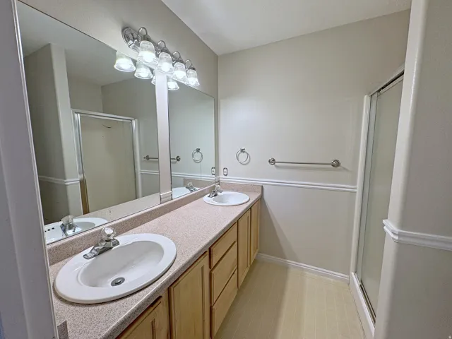 Dual vanity with integrated sinks, expansive mirror, light wood-finish cabinetry, and a walk-in shower with sliding glass door