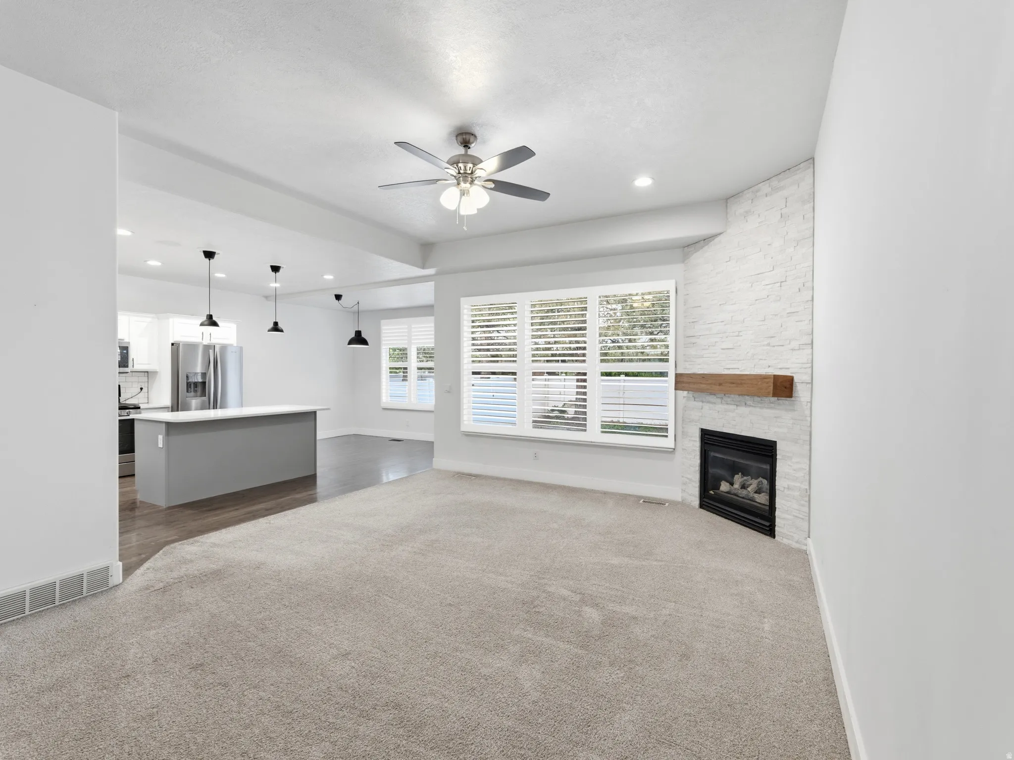 Living area with neutral carpeting, a fireplace featuring a white brick-finish surround and wood mantel, and multiple windows with plantation shutters