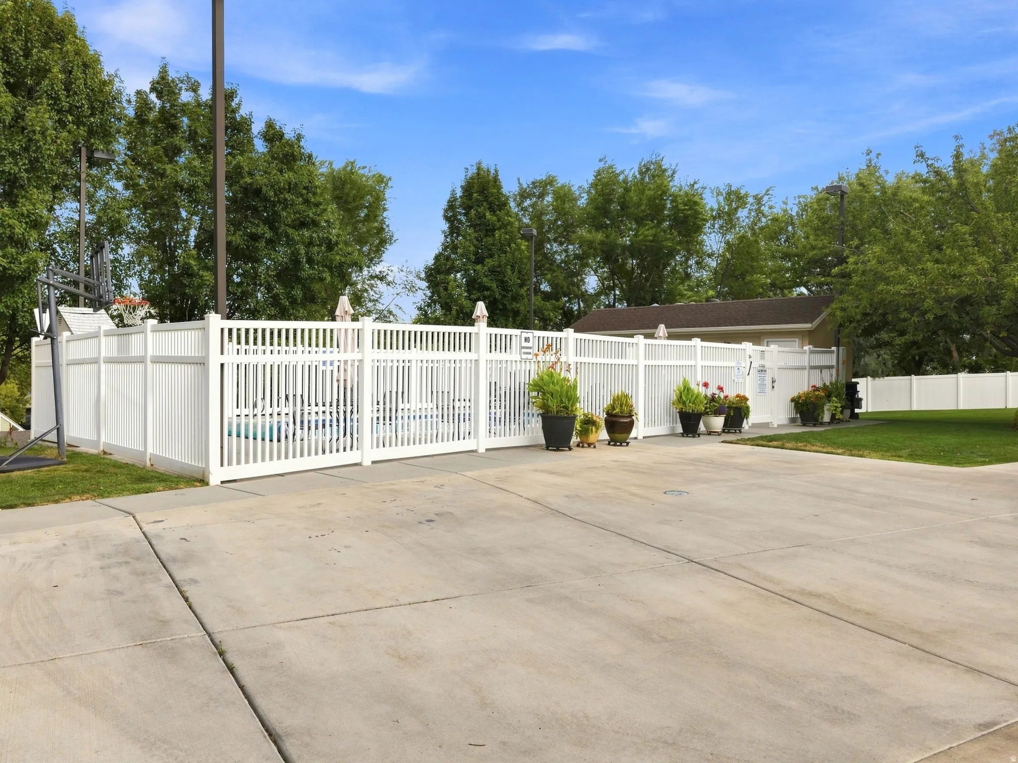 Paved concrete surface with a white picket fence enclosing a pool area, featuring mature trees and a structured building in the background