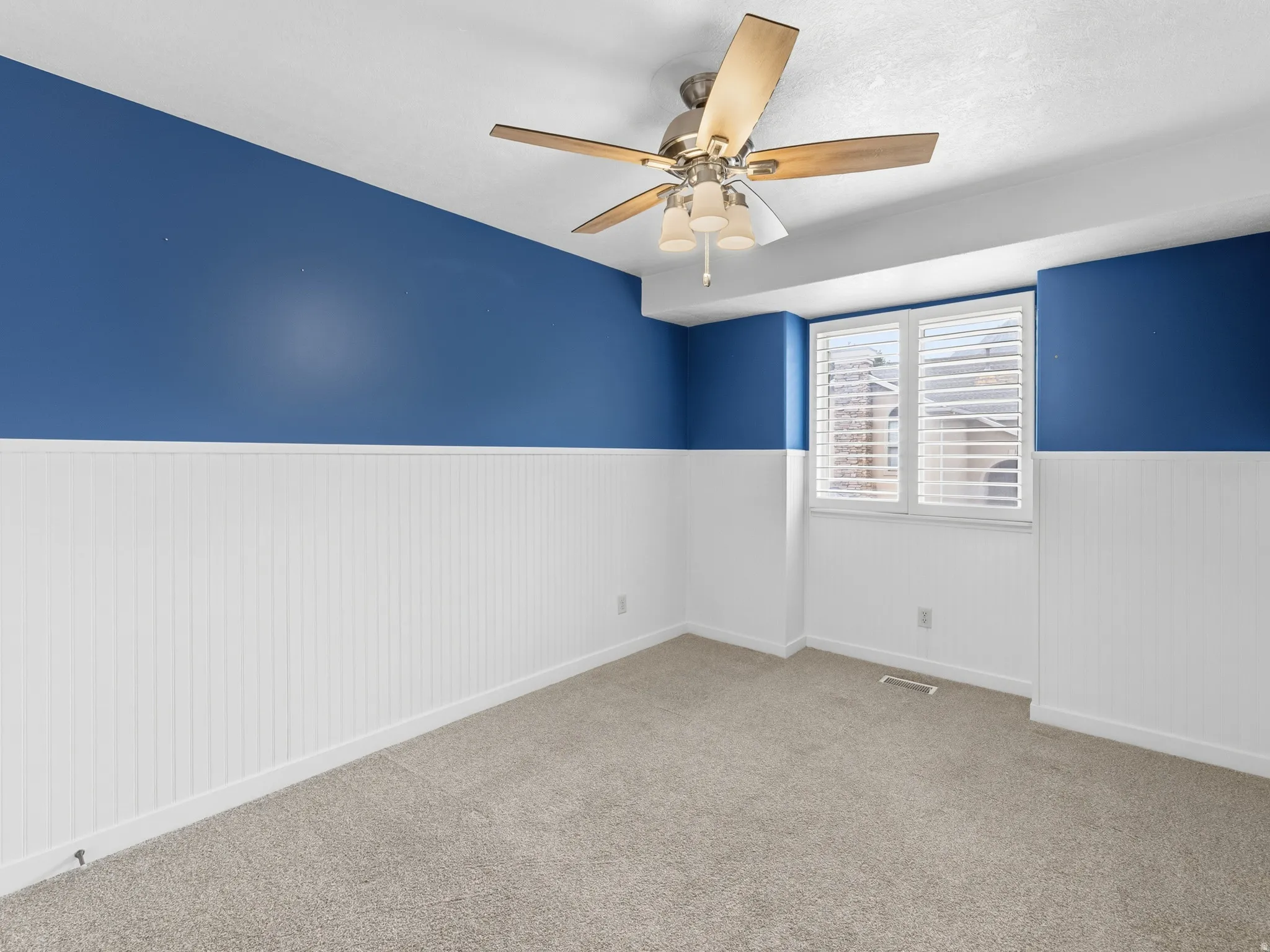 Carpeted room featuring a window with plantation shutters, a ceiling fan with integrated lighting, and walls with beadboard paneling