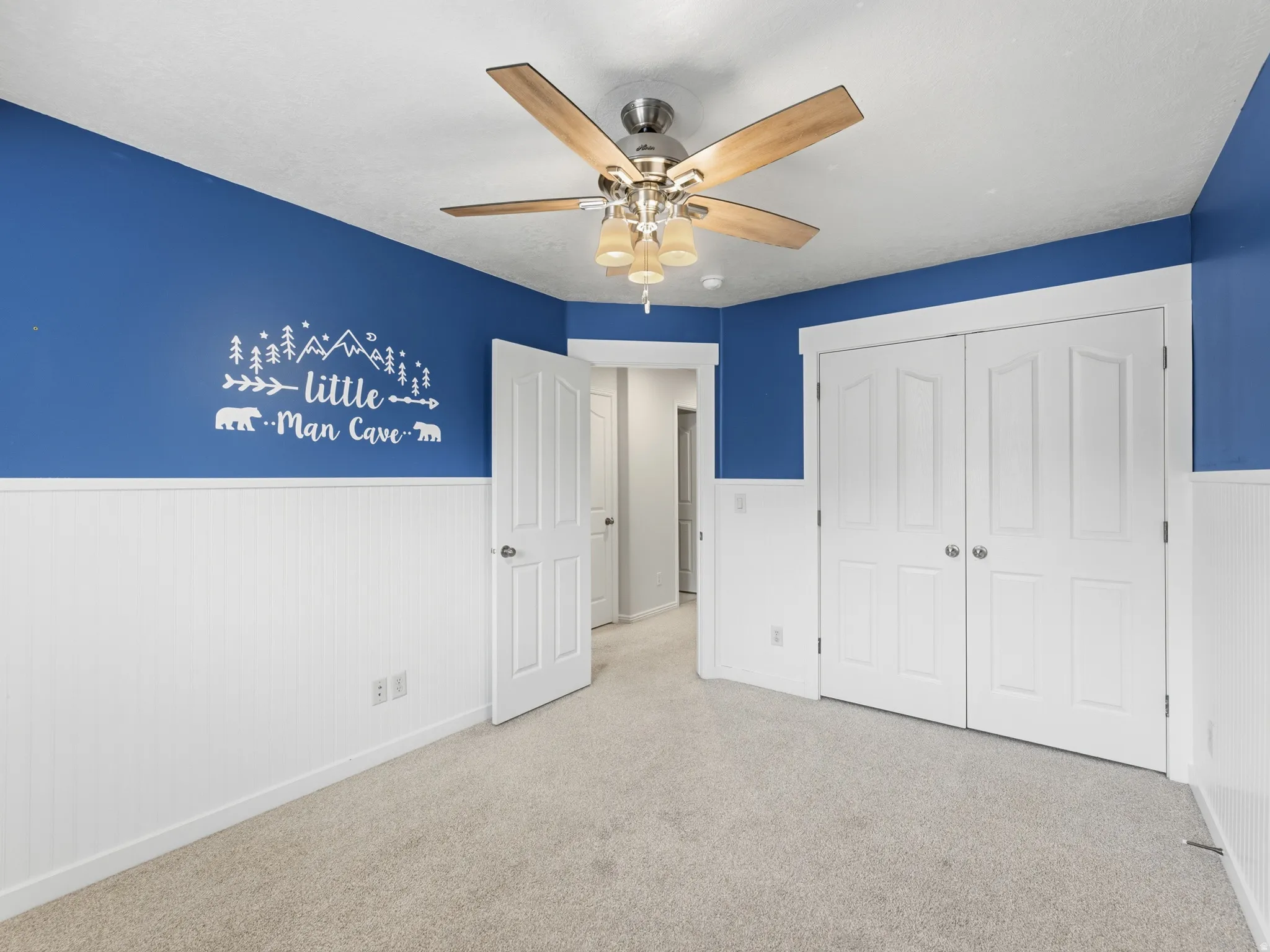White beadboard wainscoting, light carpet flooring, ceiling fan with light fixture, and white bi-fold closet doors