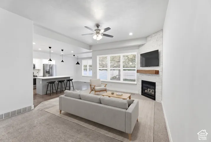 Living area featuring a fireplace with a wood mantle, large windows with blinds, and a ceiling fan