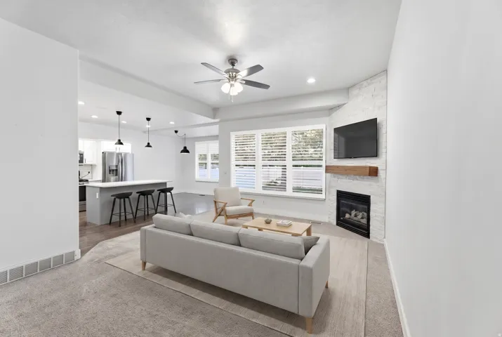 Living area featuring a fireplace with a wood mantle, large windows with blinds, and a ceiling fan