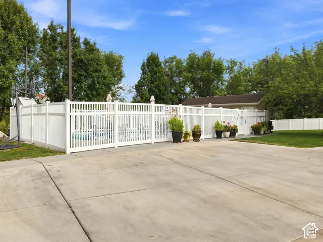 Paved concrete surface with a white picket fence enclosing a pool area, featuring mature trees and a structured building in the background