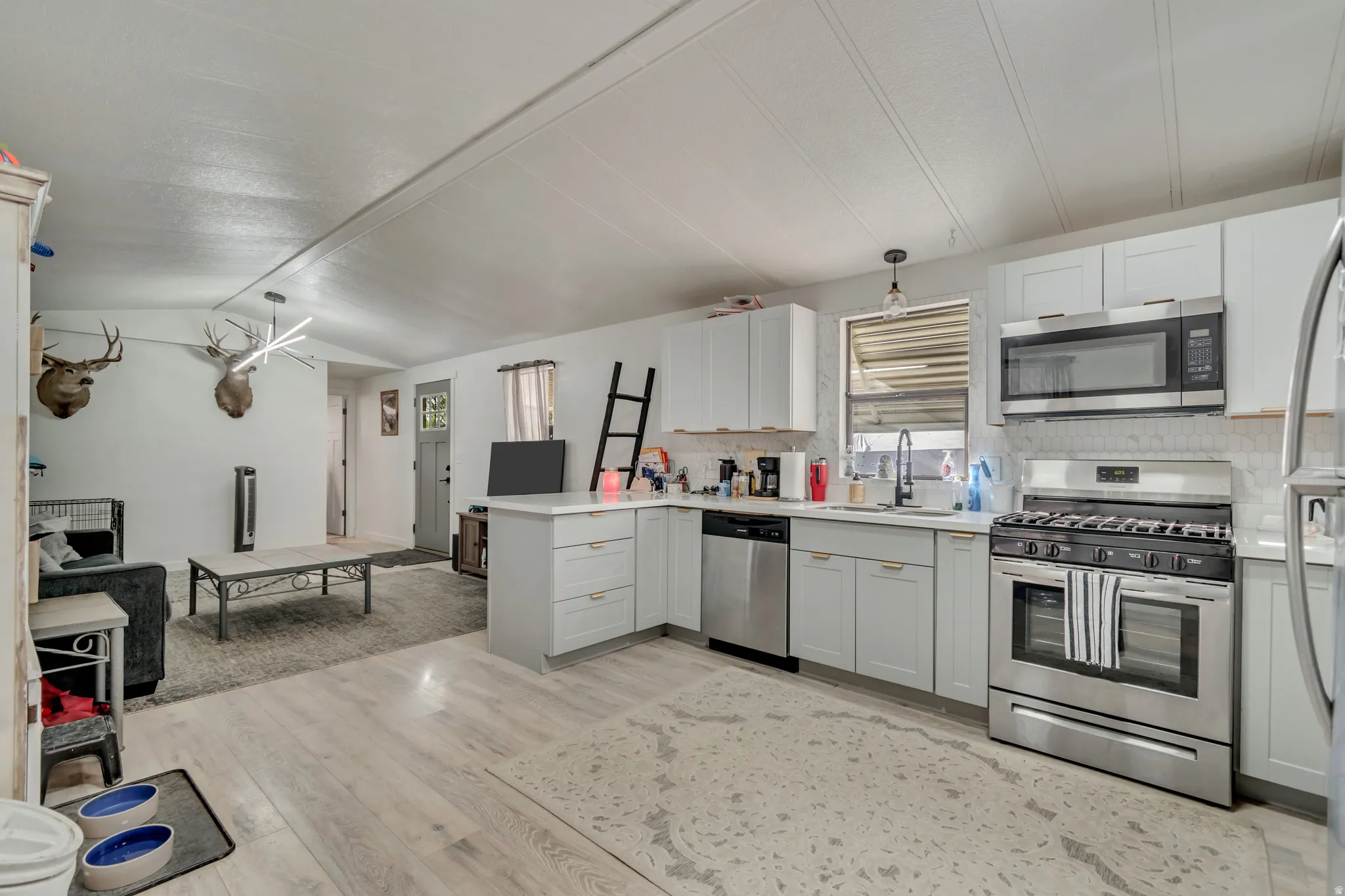 Open-concept kitchen and living area featuring wood-finish flooring, light gray cabinetry with gold-tone hardware, stainless steel appliances, and white countertops