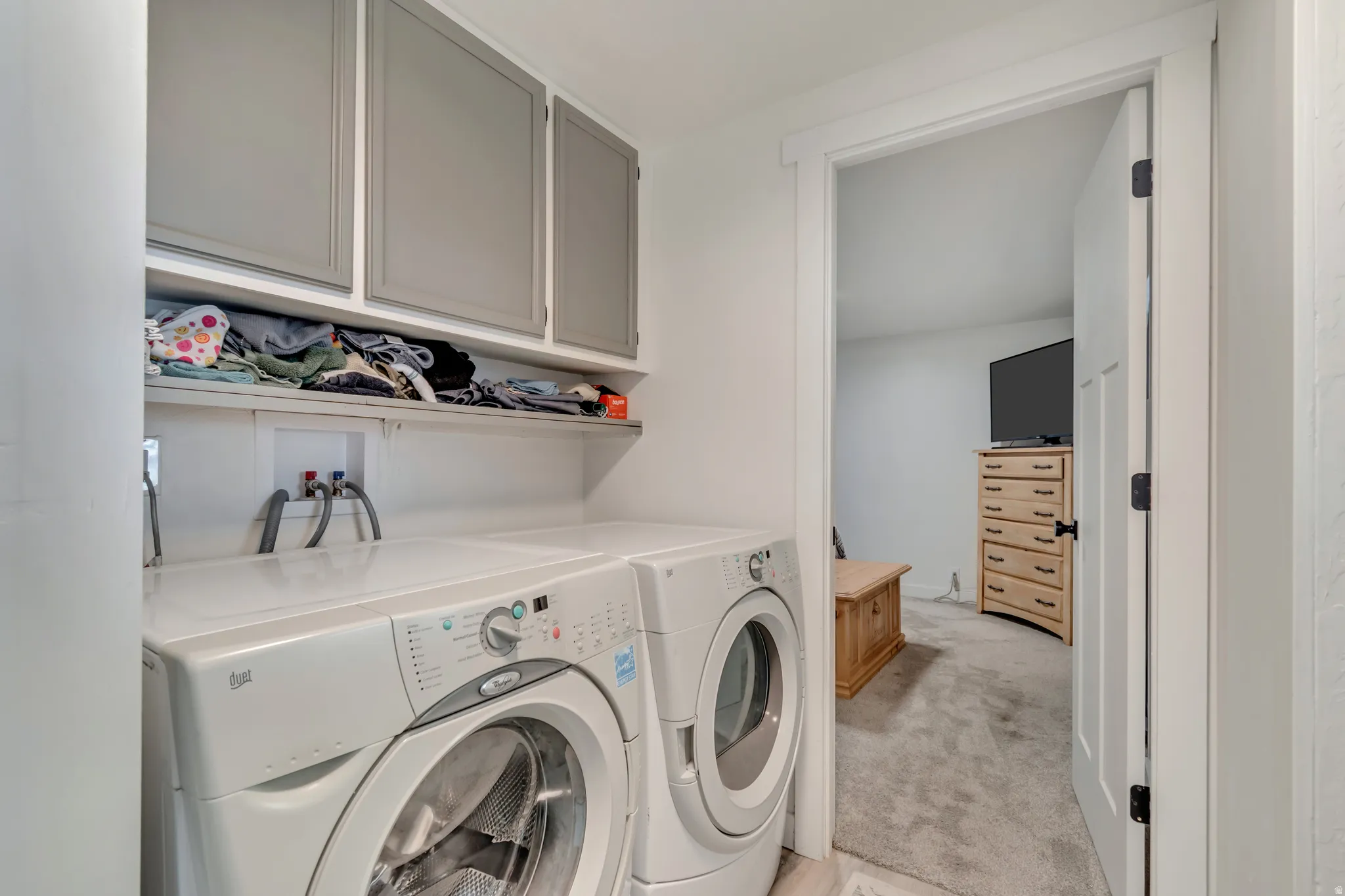 Dedicated laundry room featuring side-by-side front-load washers and dryers, white cabinetry with frosted-glass upper cabinets, quartz countertop, and built-in shelving for storage and supplies