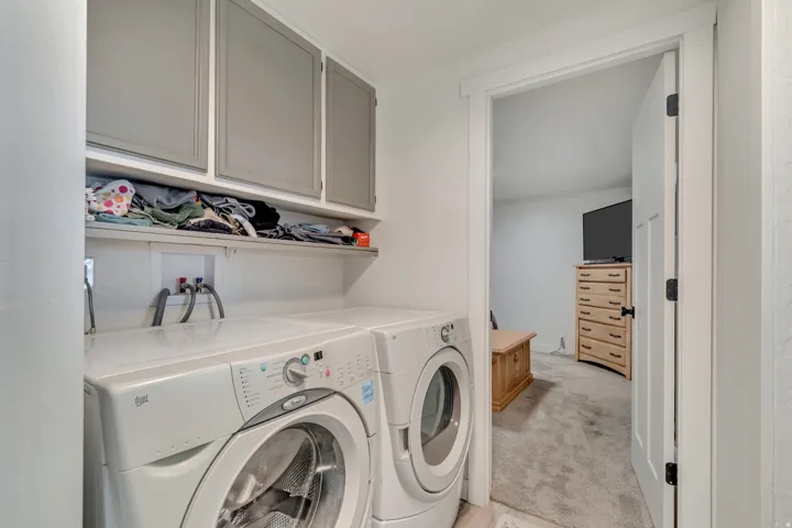 Dedicated laundry room featuring side-by-side front-load washers and dryers, white cabinetry with frosted-glass upper cabinets, quartz countertop, and built-in shelving for storage and supplies