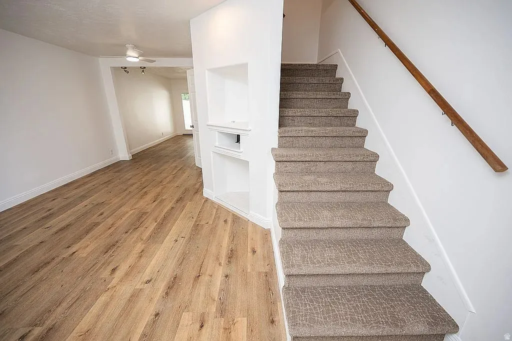 Wood-finish flooring throughout, built-in shelving, carpeted staircase, and an overhead ceiling fan