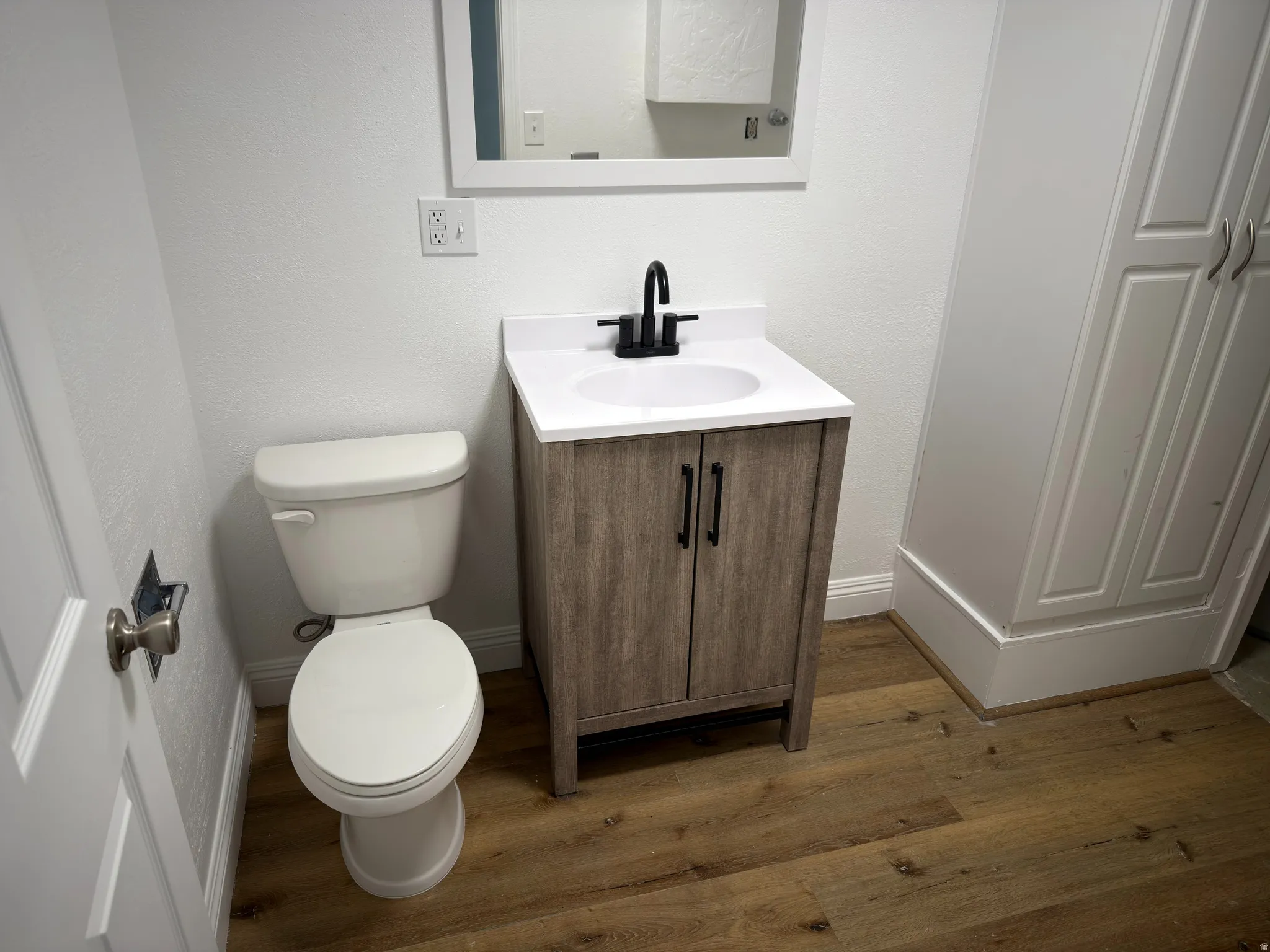 Wood-finish flooring, white vanity with black hardware, white countertop with integrated sink, wall-mounted mirror, and white built-in cabinetry