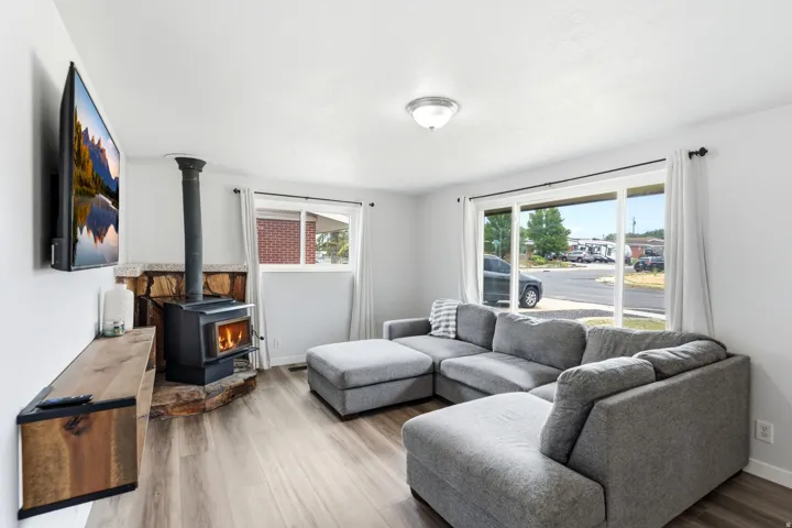 Bright, open living area showcasing wood-finish flooring, expansive windows with natural light, and a wood-burning stove with black chimney