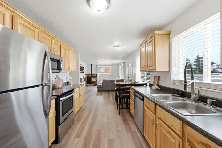 Galley kitchen featuring stainless steel appliances, natural wood cabinetry, black countertops, stainless steel sinks, and wood-finish flooring throughout