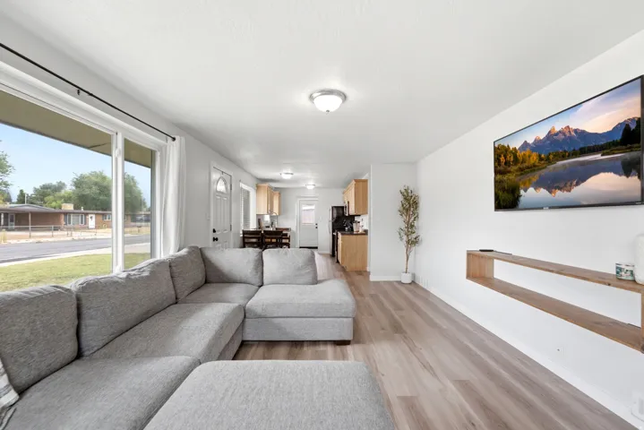 Bright open-concept living space featuring a gray upholstered sectional sofa, wood-finish flooring throughout, and expansive windows overlooking the front landscape