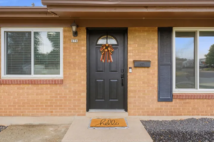 Welcoming entry featuring a brick facade, black six-panel entry door with transom window and decorative hardware, flanked by large windows with integrated blinds