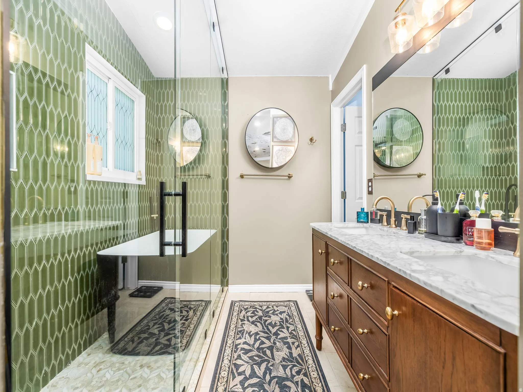 Walk-in shower with green geometric tile, freestanding bathtub, dual vanity with wood cabinetry, marble countertop, and brass fixtures