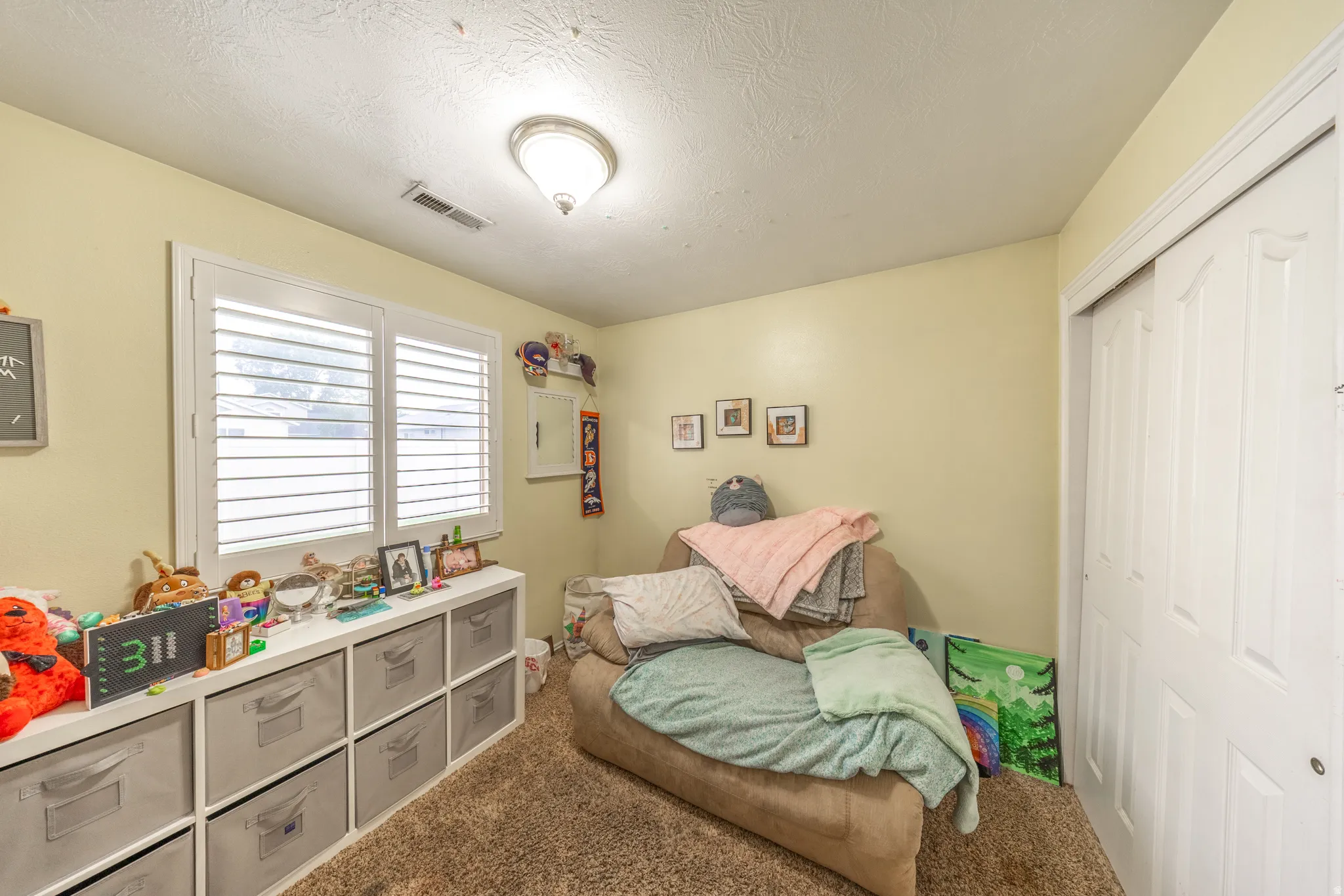 Bedroom #2 features soft carpeting, a wall-mounted light fixture, and a window accented with classic plantation shutters.