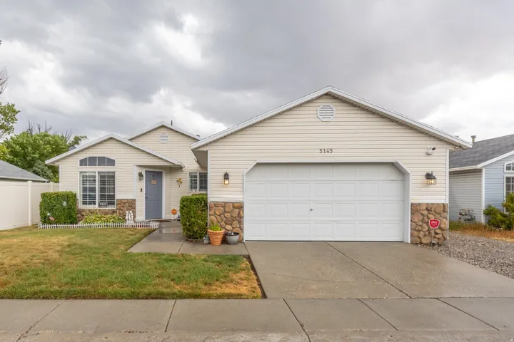 Charming exterior featuring light-colored siding complemented by attractive stone accents, an attached garage, classic white picket fencing, and a spacious concrete driveway.