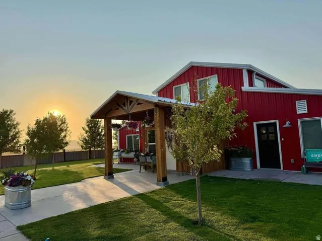 Red barn-style residence with modern updates, featuring a covered wood-beam pavilion with concrete patio, mature landscaping including trees and lawn, metal roof accents, and multiple windows with dark trim throughout the primary structure