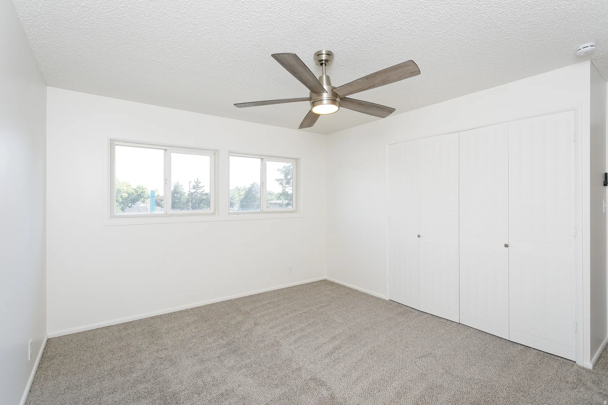 Grey carpet flooring, white walls, multiple windows, brushed nickel ceiling fan, and bi-fold closet doors